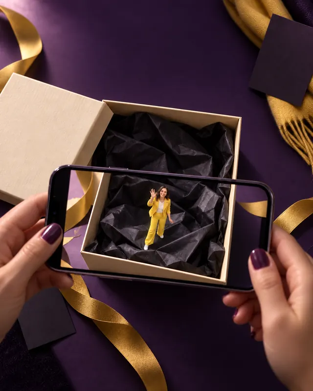 AR concept: two hands hold a phone over an open gift box with black tissue paper; on the screen, a tiny woman in a yellow suit waves from inside the box.